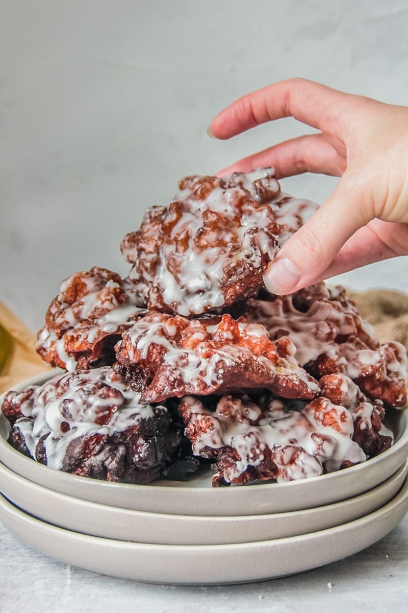 A glazed apple fritter is picked up from a stack on a stack of rimmed ceramic plates on a gray surface.