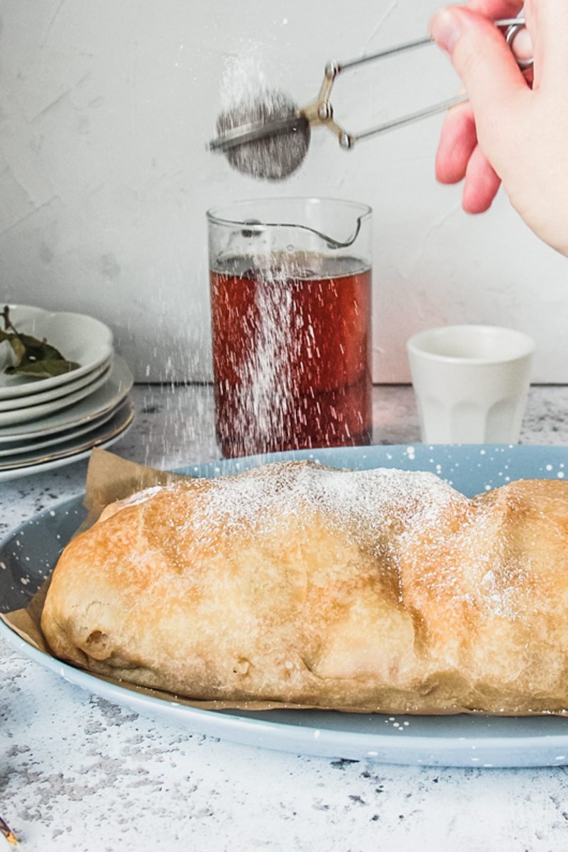 Powdered sugar is sprinkled over the top of homemade apple strudel served up on a blue plate on a light gray surface.