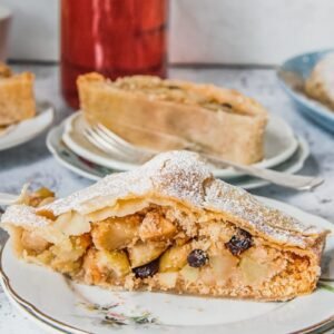 A slice of apple strudel sits on a white plates with other pieces in the background on a light gray surface.