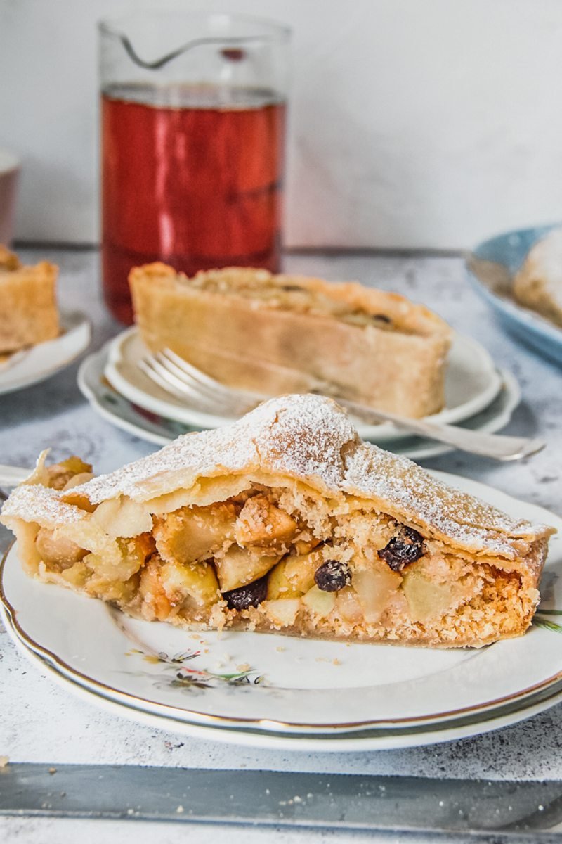 A slice of apple strudel sits on a white plates with other pieces in the background on a light gray surface.