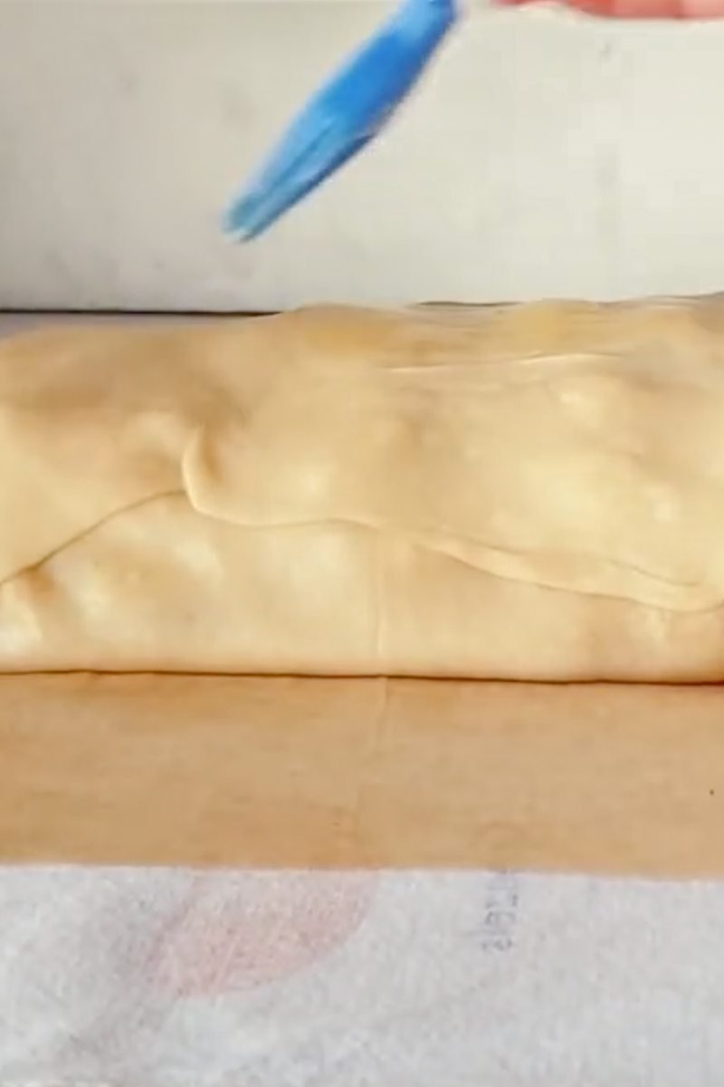 Unbaked apple strudel is brushed with melted buter on parchment paper on a light gray surface.