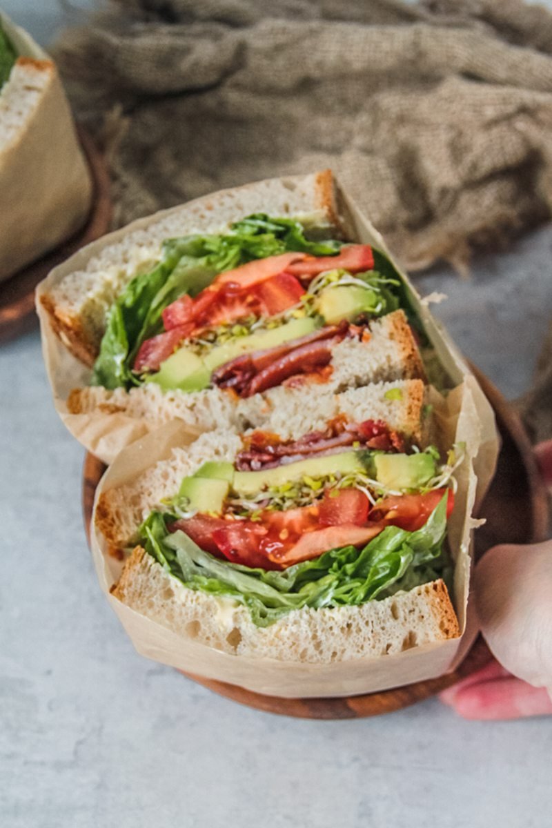 A halved BLATS sandwich sitting on a wooden plate is picked up.