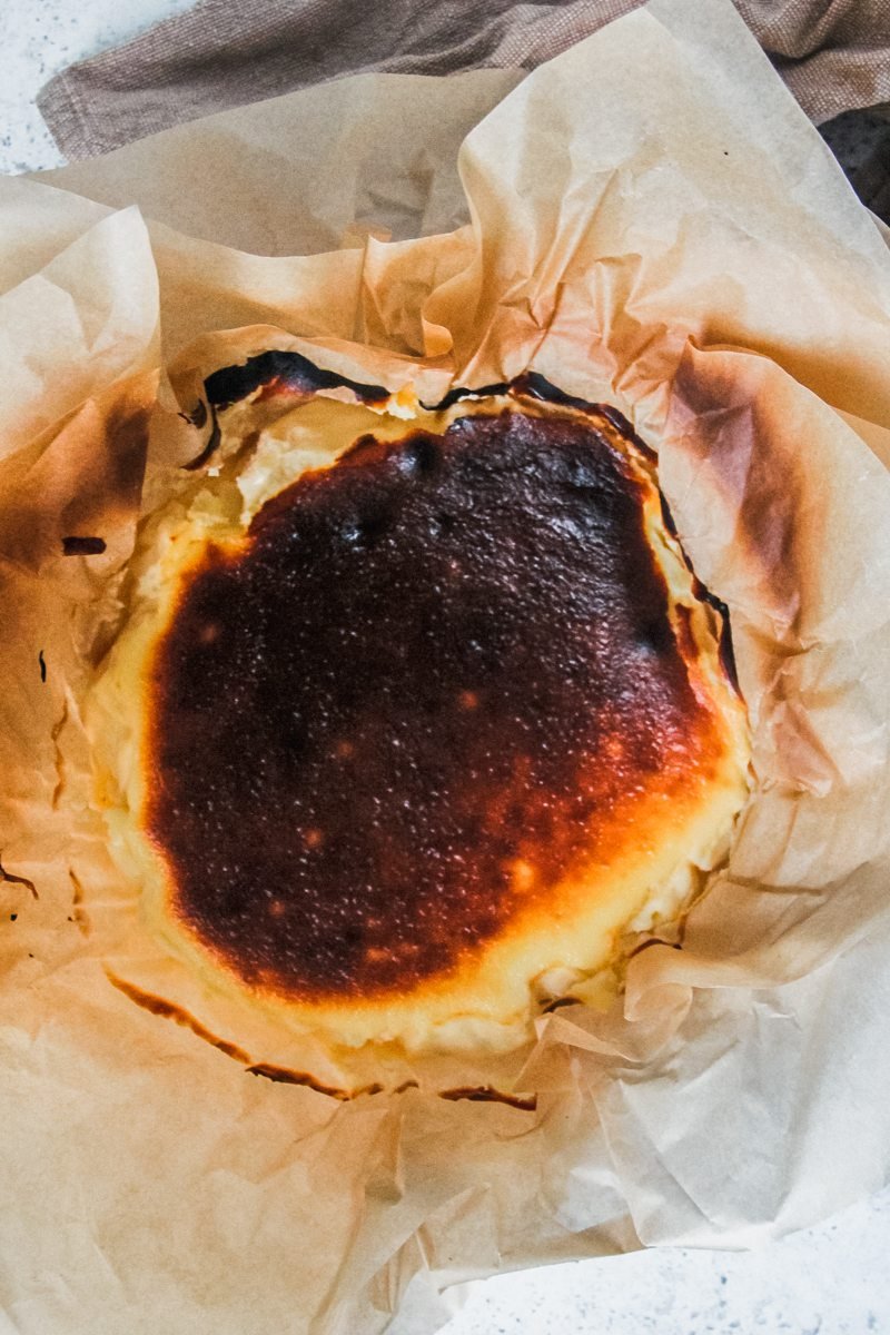 Basque burnt cheesecake sits in layers of parchment paper with it's deeply caramelized top on a light gray surface.