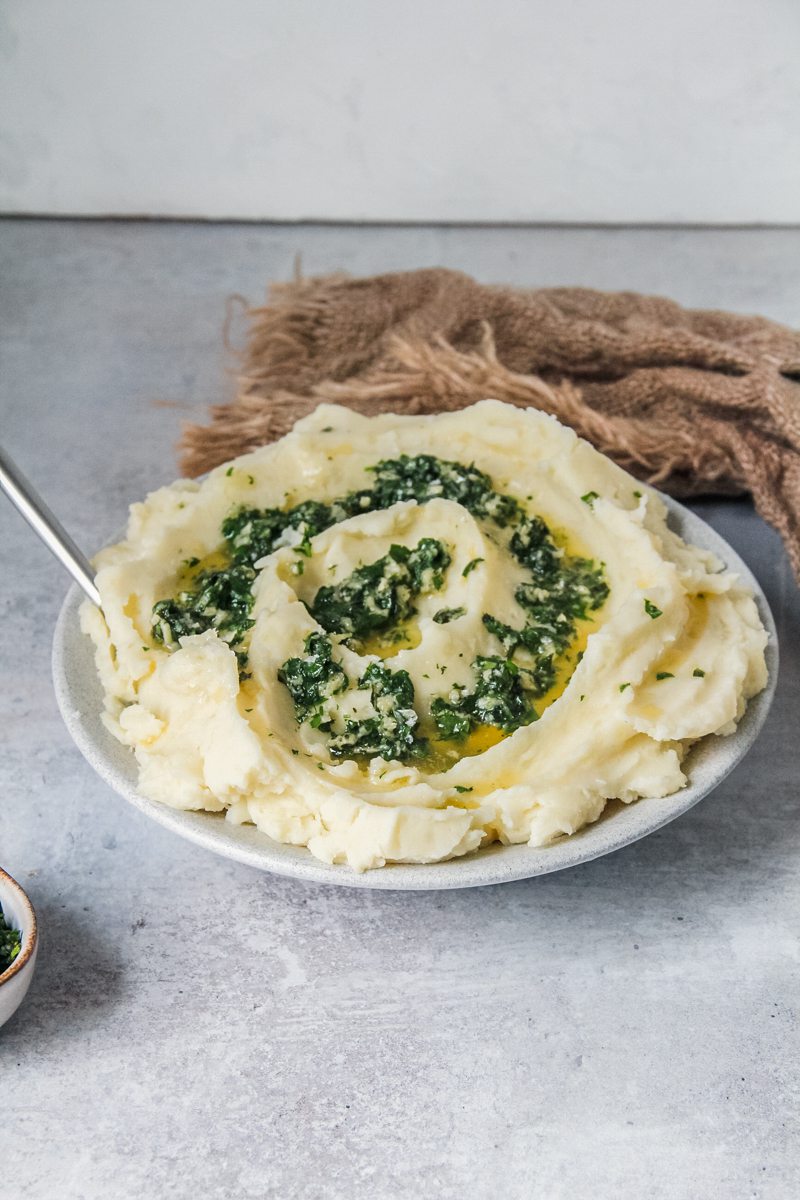 A side angle of mashed potatoes topped with a garlic herb mixture on a white plate on a gray surface.