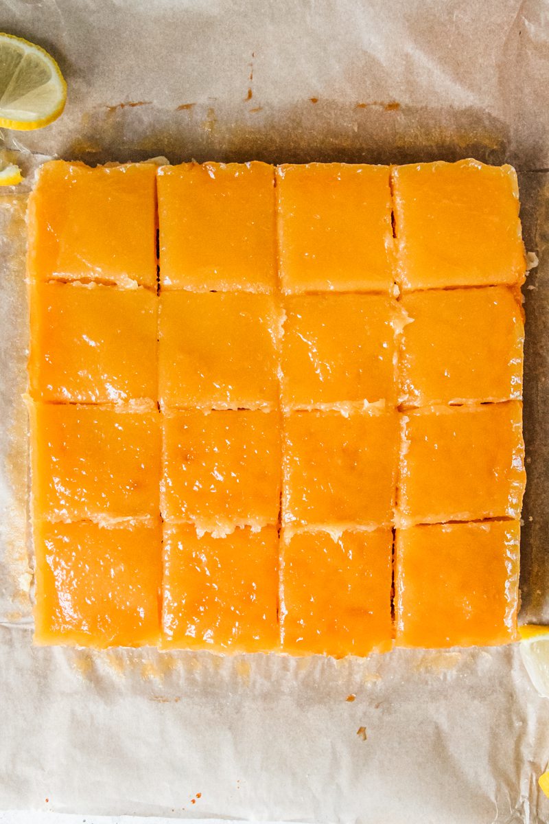 Square lemon bars sit on a brown parchment paper surface.