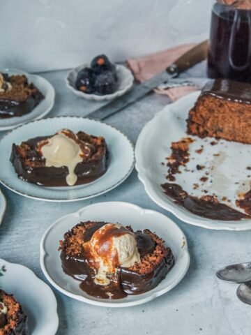 Slices of sticky toffee pudding cake sit on individual plates with a scoop of melting vanilla ice cream and toffee sauce on top on a gray surface.