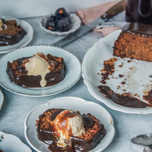 Slices of sticky toffee pudding cake sit on individual plates with a scoop of melting vanilla ice cream and toffee sauce on top on a gray surface.