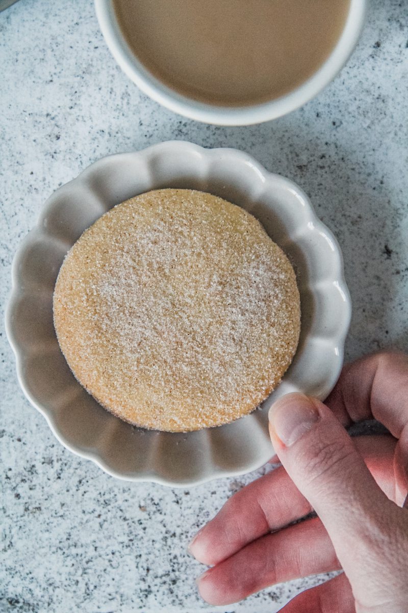 A biscochito cookie sits on a scalloped edge gray ceramic plate that is being picked up beside a cup of tea.