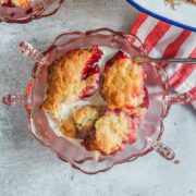 An individual serving of rhubarb and blackberry cobbler sits in pink glass bowls on a gray surface with a spoon resting and cream on top.
