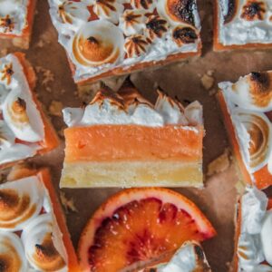 Blood orange curd shortbread bars sit on parchment paper with one on it's side showing the shortbread, curd ceenter and caramelized meringue topping.