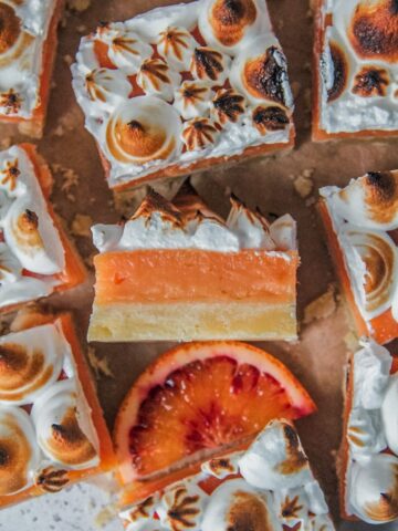 Blood orange curd shortbread bars sit on parchment paper with one on it's side showing the shortbread, curd ceenter and caramelized meringue topping.