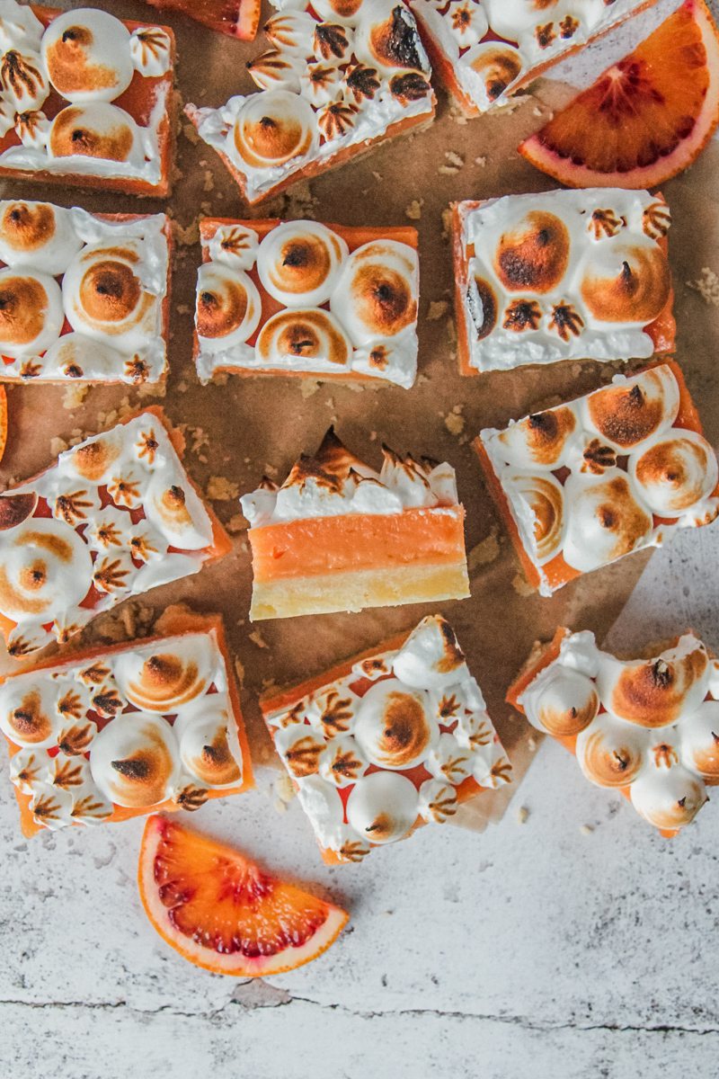 Blood orange shortbread meringue bars sit on a parchment paper lined light gray surface with one sitting on it's side showing the shortbread base and curd center and caramelized meringue layers.