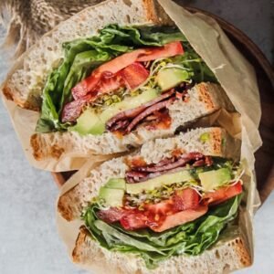 A halved BLATS sandwich sits wrapped in parchment paper laying with the filling visible, on a wooden plate on a gray surface.