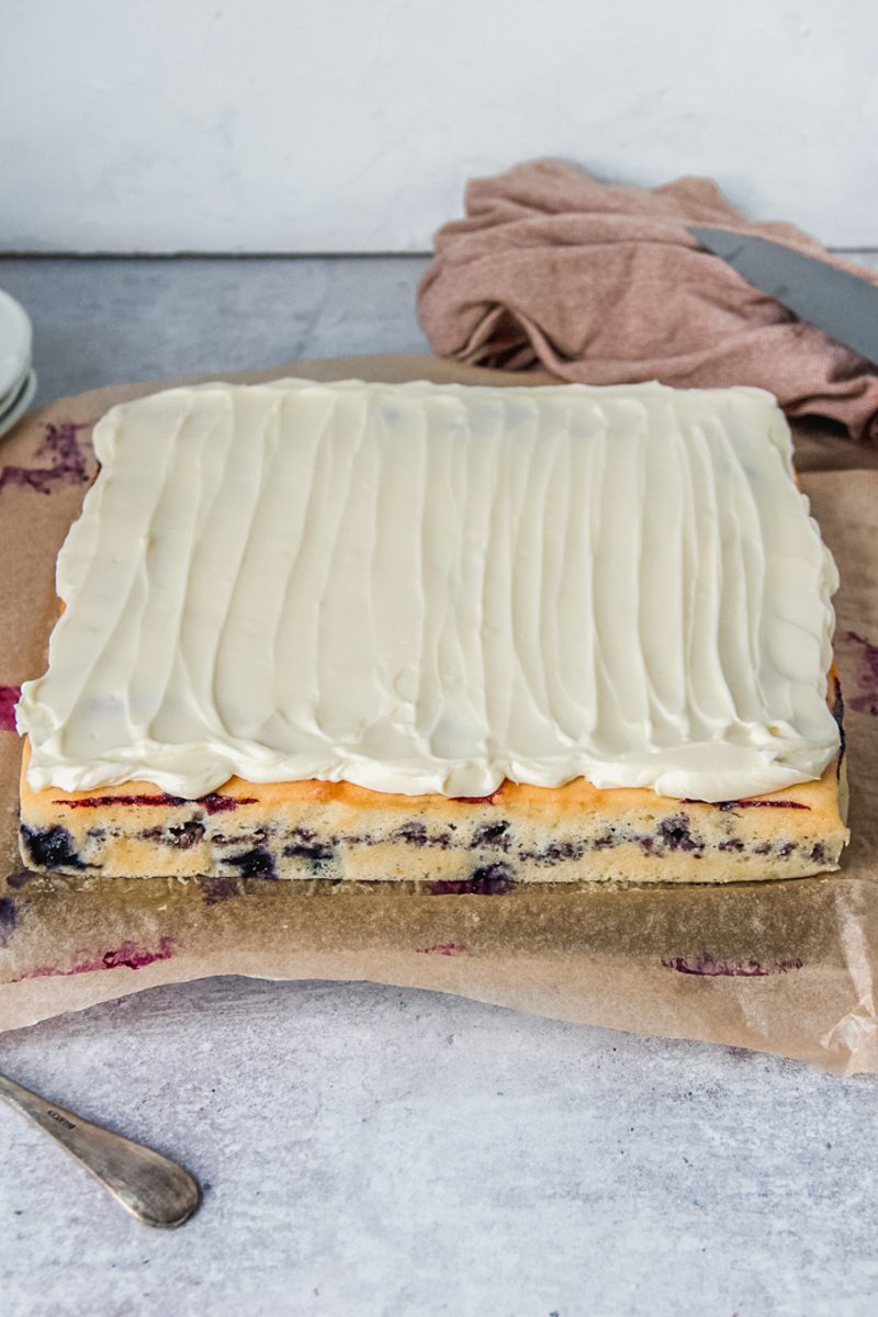 Blueberry sheet cake topped with whipped cream cheese frosting sits on brown paper on a gray surface.