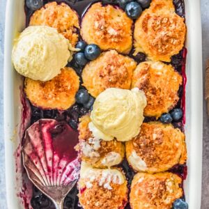 Blueberry cobbler sits with a serving spoon resting in a rectangle white ceramic baking dish on a gray surface.