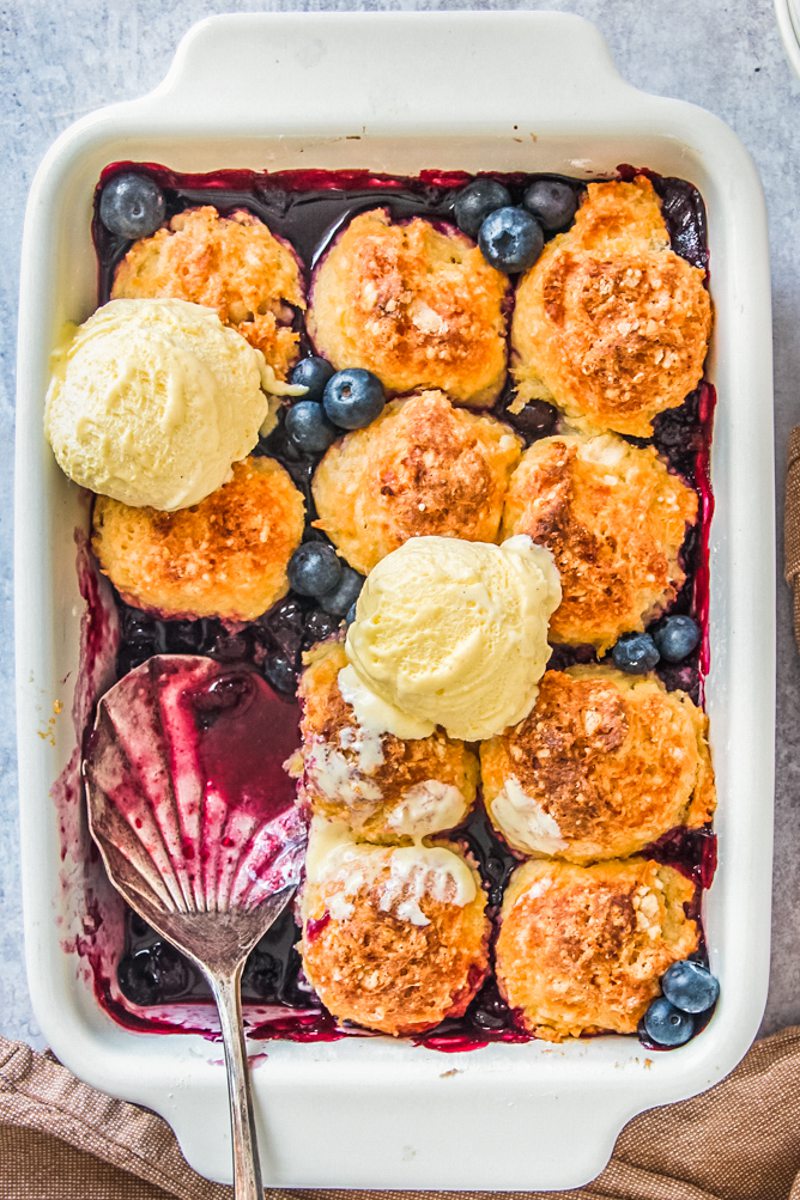 Blueberry cobbler sits with a serving spoon resting in a rectangle white ceramic baking dish on a gray surface.