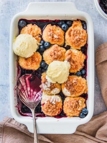 A blueberry cobbler is served up in the white rectangle baking dish it was baked in with scoops of vanilla ice cream decorating the corn dumpling topping, on a light gray background.