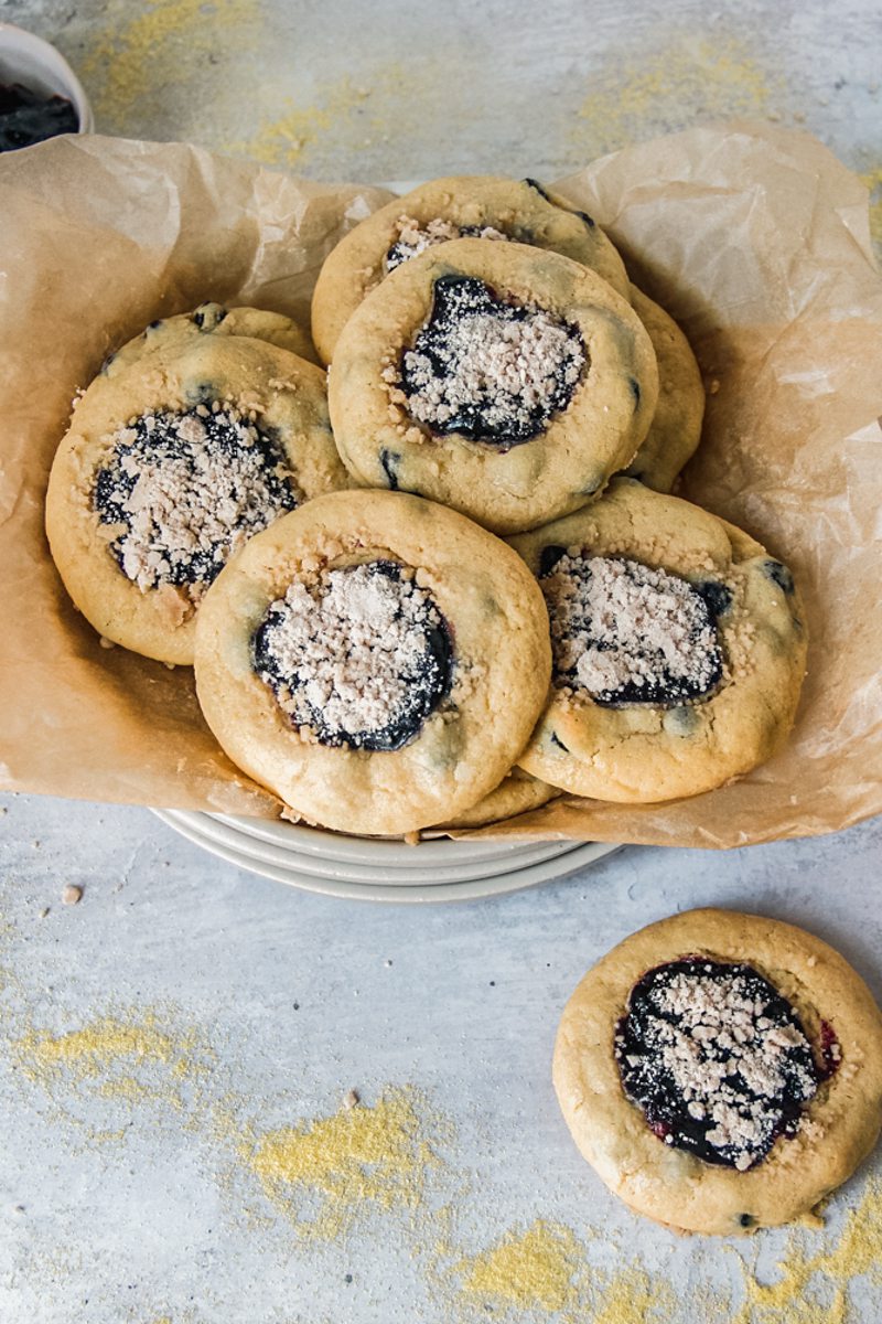 Blueberry muffin cookies sit roughly stacked on a parchment lined stack of ceramic plates on a gray surface.