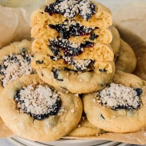 Halved blueberry muffin cookies sit stacked on a parchment lined stack of ceramic plates on a gray surface.