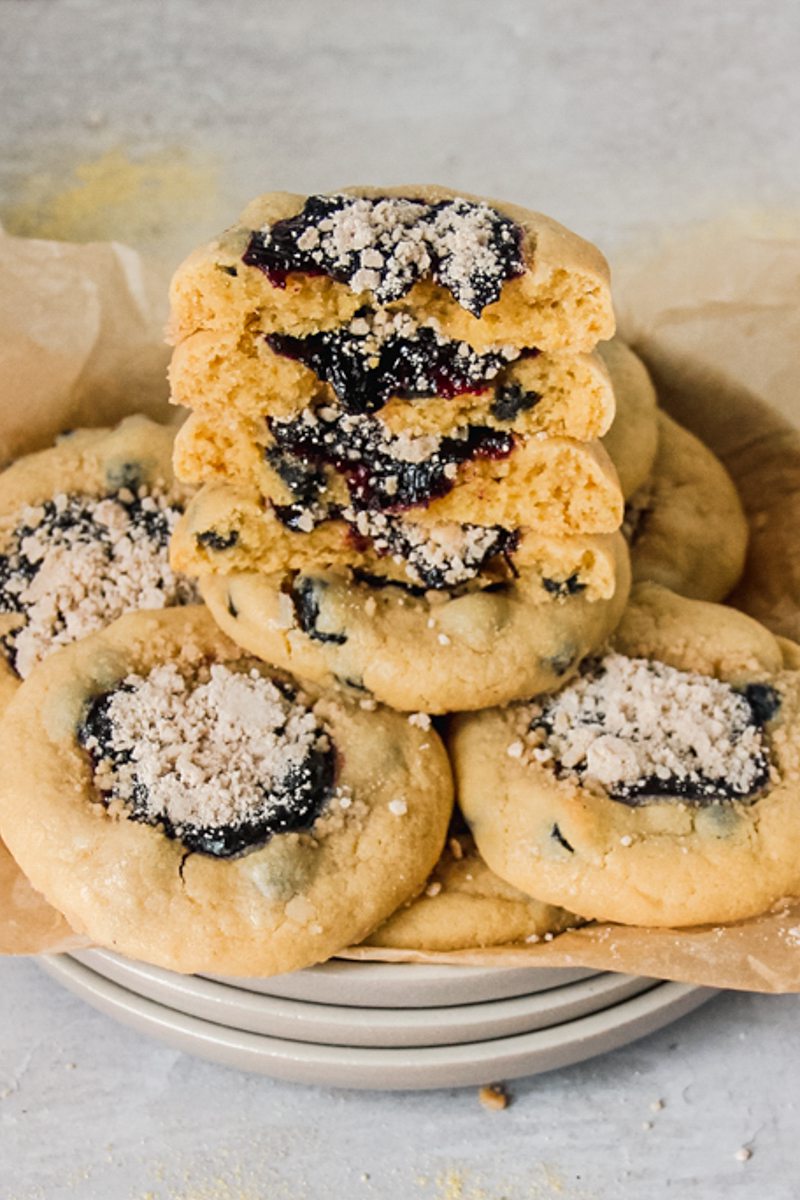 Halved blueberry muffin cookies sit stacked on a parchment lined stack of ceramic plates on a gray surface.