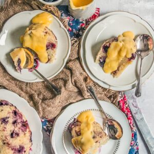 Individual servings of blueberry duff steamed pudding with thick drizzles of custard sit on white ceramic plates on a brown cloth on a gray surface.