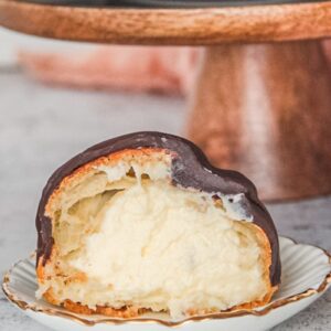 A halved Dutch profiterole sits on an individual plate filled with whipped cream with a chocolate topping on a light gray surface.