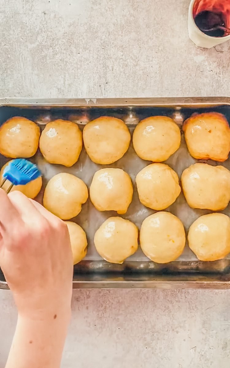 Rolls of filled dough are brushed with melted butter in a stainless steel baking tin on a gray surface.