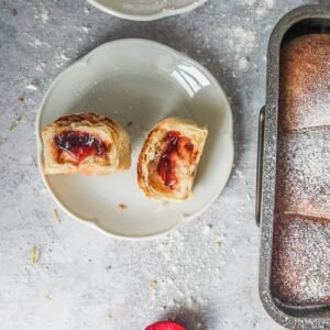 A European jam filled bread roll sits torn in half on a white plate on a gray surface with another roll served up and the tin of bread rolls beside.