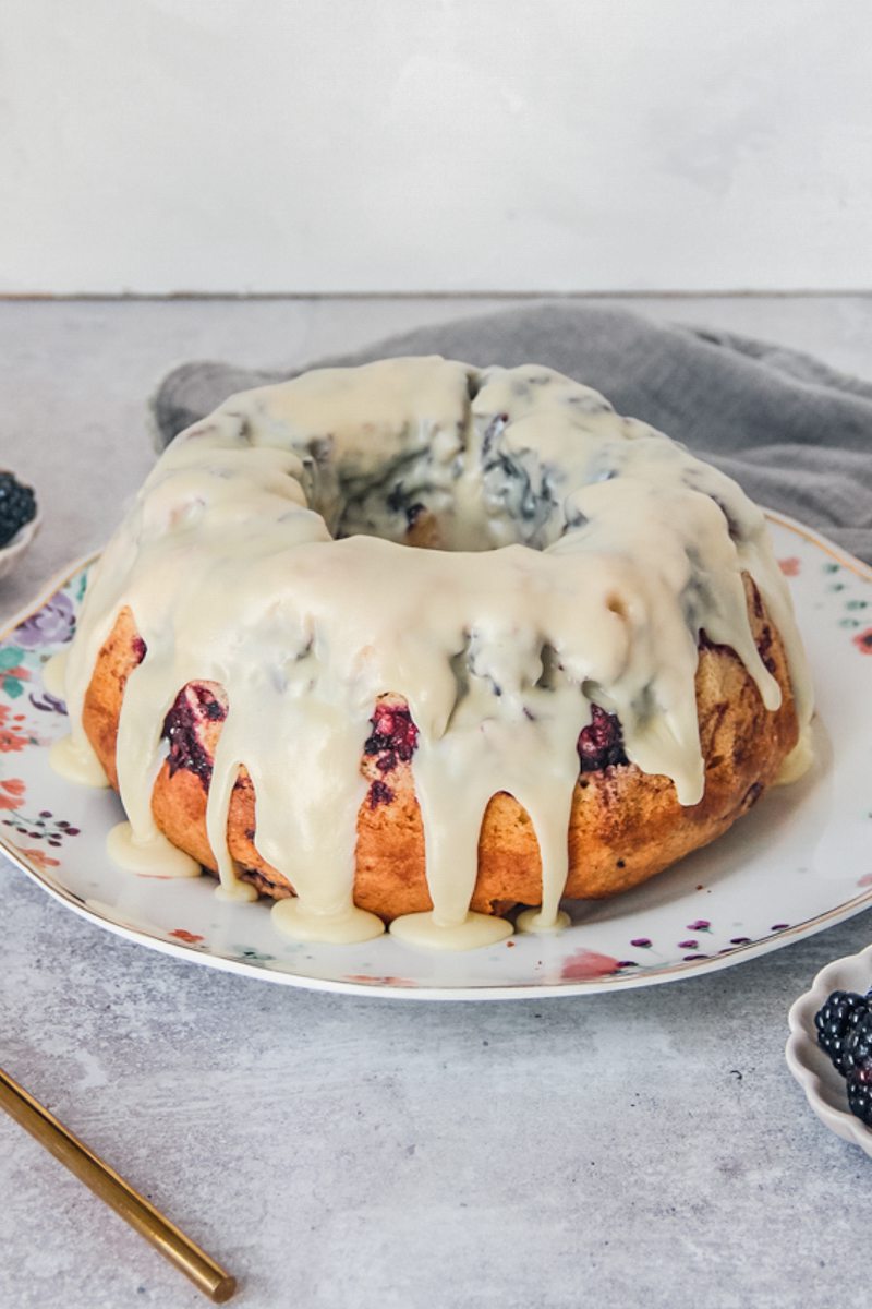 A white chocolate glaze slides over the edges of a white chocolate blackberry bundt cake on a floral ceramic plate on a gray surface.
