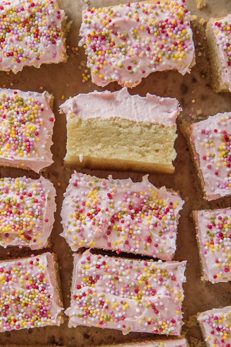 Sugar cookie bars sit on a parchment paper with a raspberry buttercream frosting and hundreds and thousands sprinkles and one on it's side with the soft sugar cookie bar and fluffy frosting visible.