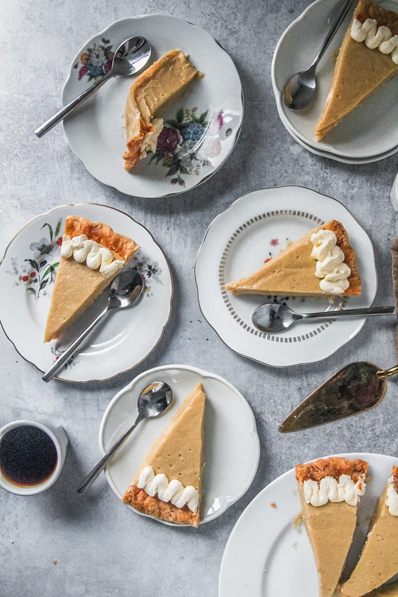 Slices of butterscotch pie sit on white ceramic plates on a gray surface with a cup of coffee beside.