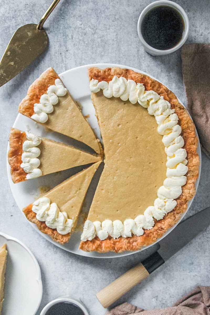 A butterscotch pie sits on a white ceramic plate on a gray surface with cups of coffee beside.