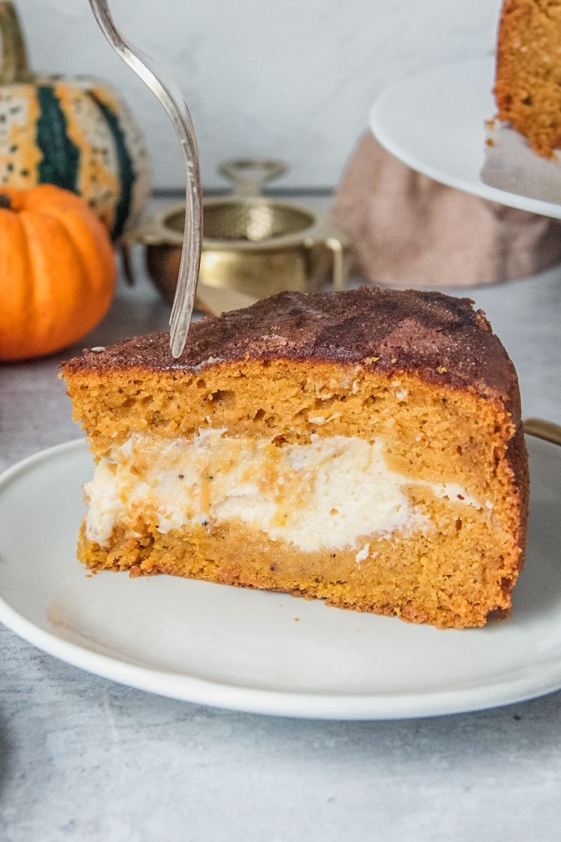 A slice of pumpkin cake is served up on a white plate with the cream cheese center visible and a fork about to be stuck into the slice.