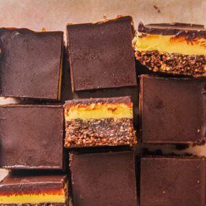 Nanaimo bars sit on a lined wooden board on a gray surface with some turned sideways to show the three layers.