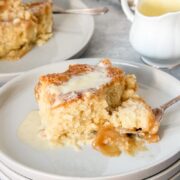 A serving of Canadian Maple Pudding sits with a spoon on a stack of rimmed ceramic plates on a gray surface.