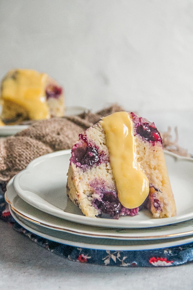 An individual serving of blueberry duff steamed pudding with a thick drizzle of custard sits on a stack of plates on a blue floral cloth with a brown cloth behind.