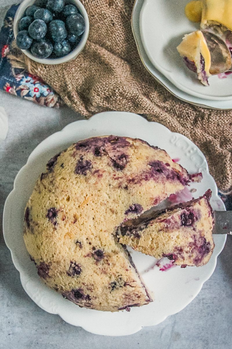 A slice of steamed blueberry pudding sits beside the remaining pudding on a white scalloped ceramic plate on a brown cloth on a gray surface.