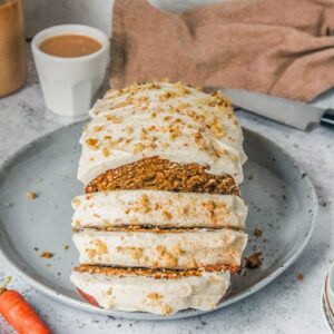 A carrot cake loaf topped with cream cheese frosting with a few plates resting against the edge of a round blue speckled ceramic plate on a light gray surface.