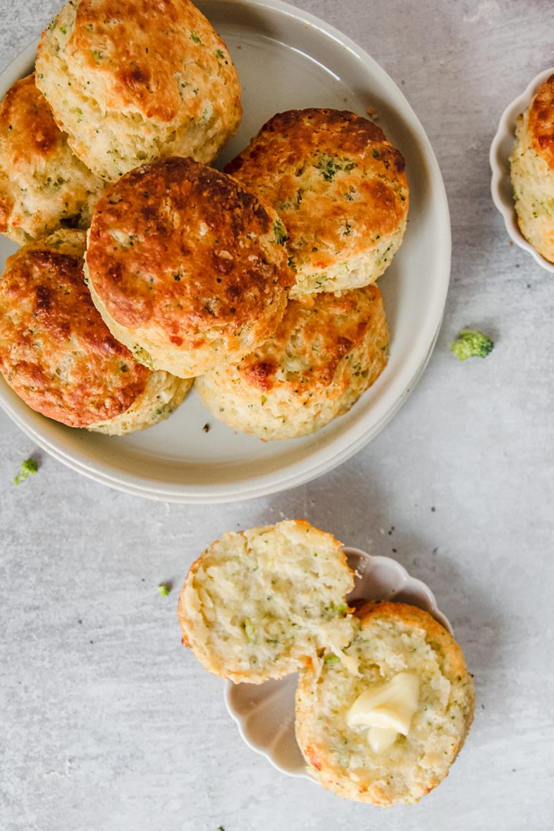 A halved savory biscuit sits on a small individual ceramic plate with a little butter in the center whilst a rimmed ceramic plate on a gray surface.