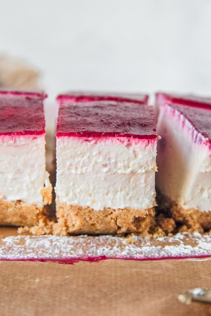 Squares of cranberry cheesecake with a crushed biscuit base, no bake filling and layer of cranberry puree sit on parchment paper.