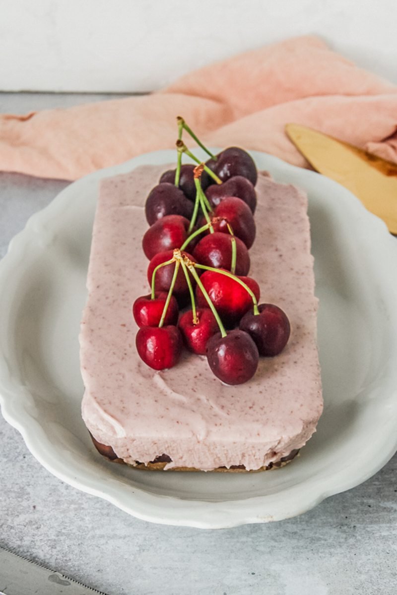 A cherry cheesecake sits on an oval white ceramic plate on a gray surface with a coral cloth.