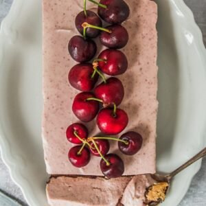 A cherry cheesecake sits on an oval white ceramic plate with a slice cut out with a spoonful of the cake resting on the plate on a gray surface.