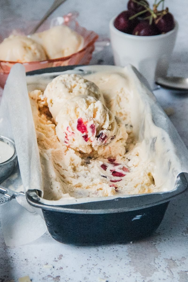 Scoops of no churn cherry ice cream semifreddo sit in a metal loaf tin on a light grey background with a pink glass bowl with a serving of the semifreddo behind the tin.