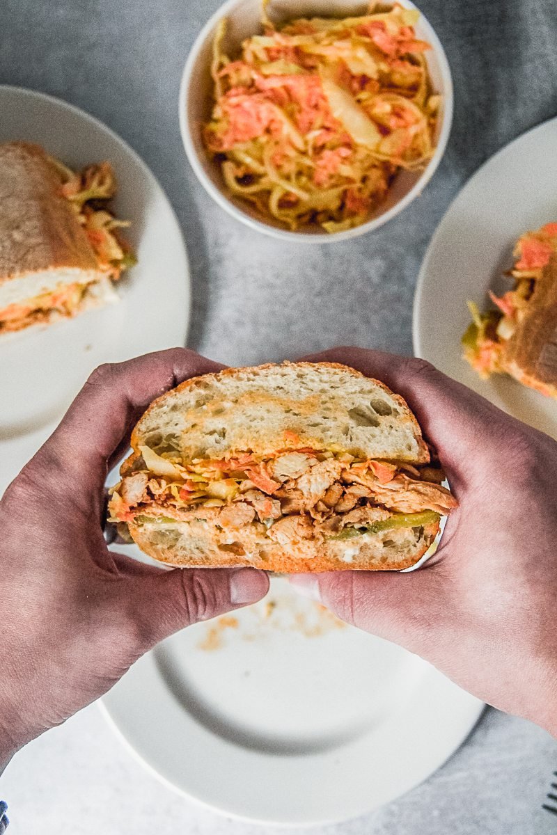 A spiced chicken and coleslaw sandwich is held with other sandwiches in the background on individual white ceramic plates and more coleslaw.