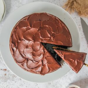 A chocolate buttermilk cake sits on a white ceramic plate with a slice being served up.