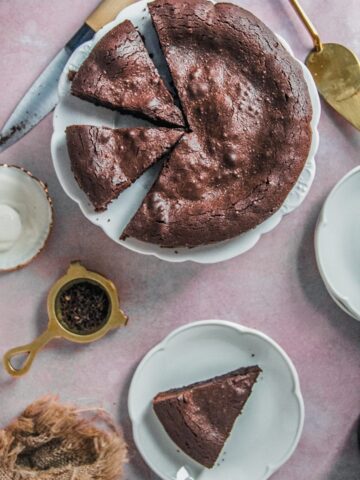 A French chocolate cake sits on a white fluted plate on a light pink background with a serving on a white plate.