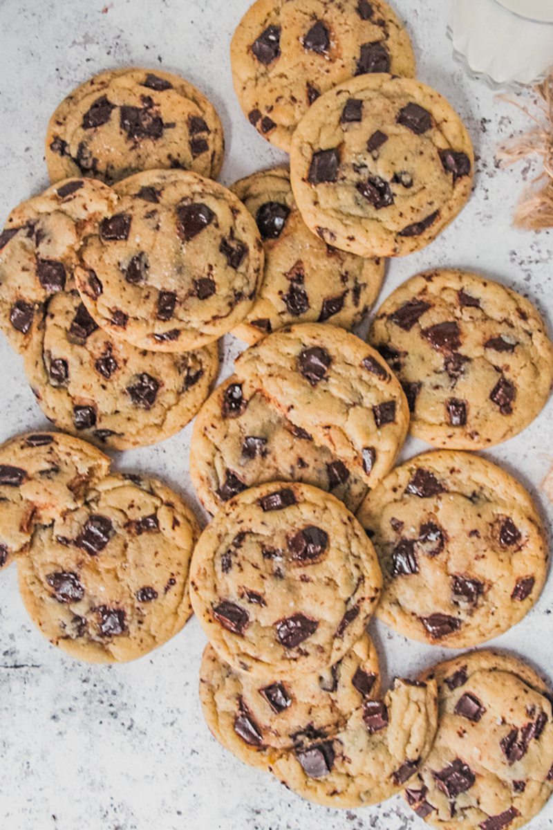 Lightly golden chocolate chunk cookies lay strewn on each other on a light gray surface.