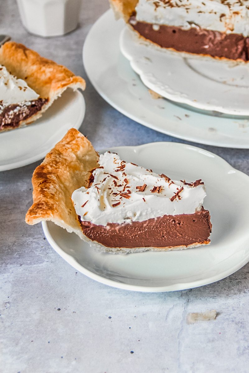 A slice of chocolate pudding pie sits on an individual white ceramic plate with a second behind and remaining pie in the background.