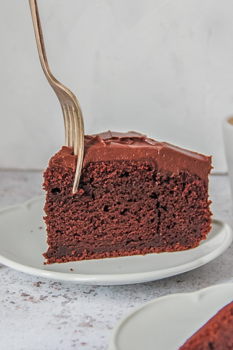 A slice of chocolate buttermilk cake sits on a white ceramic plate on a light gray surface with a fork piercing the front of the cake.