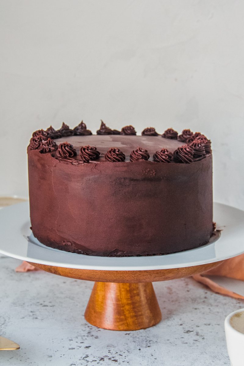 A chocolate buttermilk cake stands on a cake stand on a light gray surface.